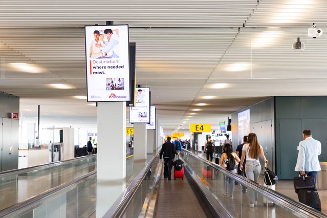 Een foto van een hal op vliegveld Schiphol waar naast de rol-paden op digitale schermen beelden van Artsen zonder Grenzen te zien zijn.